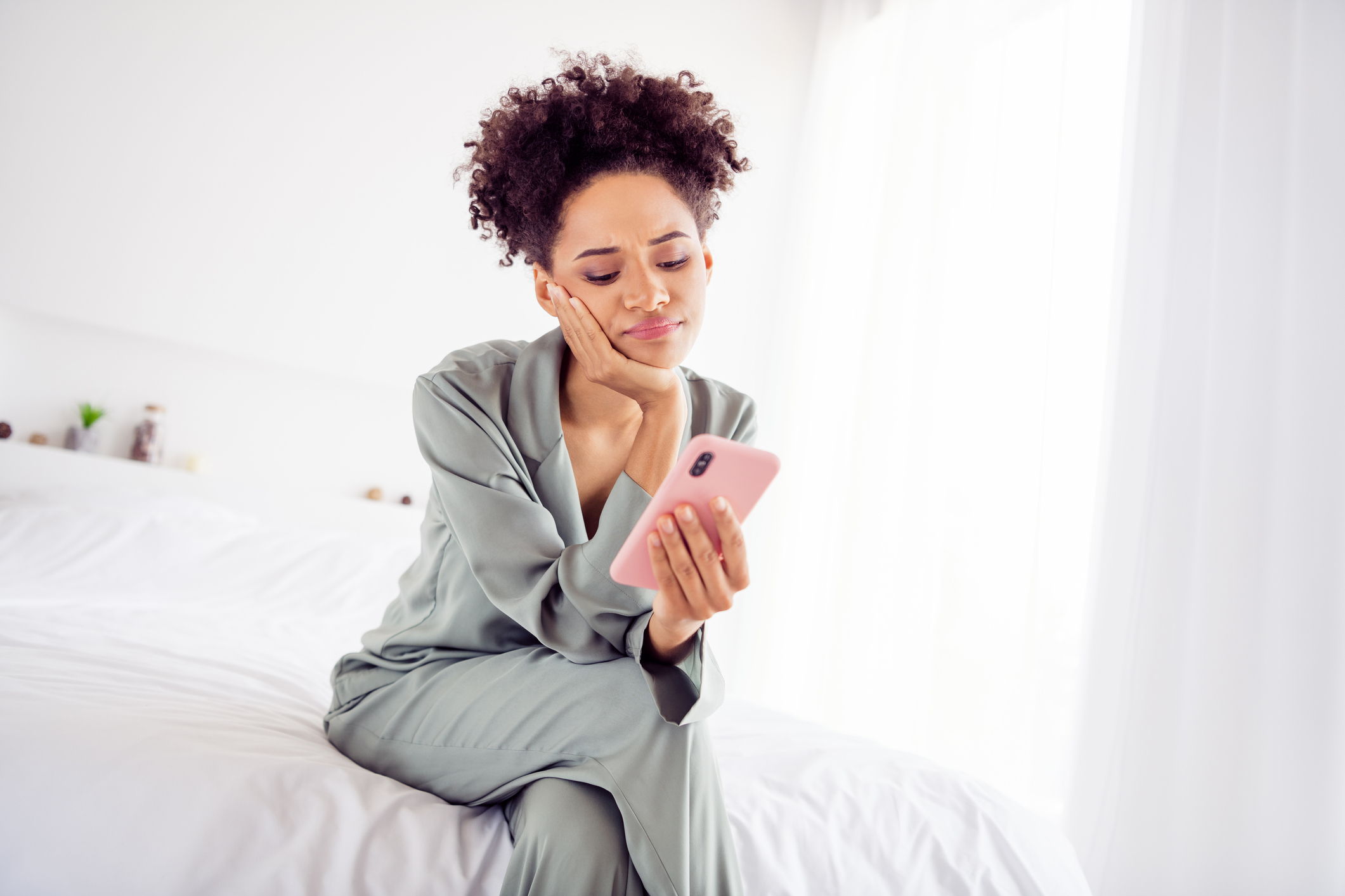 African American woman sitting on edge of bed in pajamas with mobile phone