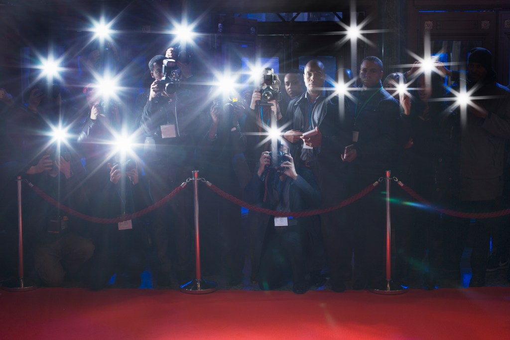 A crowd of photographers with cameras and flashing lights at a red carpet event.